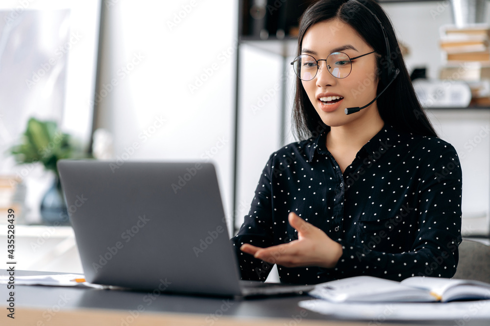 Confident pretty smart asian girl with glasses and headphones, freelancer, call center operator, sitting at desk in office, talks to client or employee via video communication, gesturing with her hand Confident pretty smart asian girl with glasses and headphones, freelancer, call center operator, sitting at desk in office, talks to client or employee via video communication, gesturing with her hand