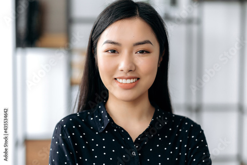 Close-up portrait of pretty happy confident asian girl. Japanese brunette young woman, wearing stylish black shirt, stand against blurred office background, looks directly at camera, smiles friendly