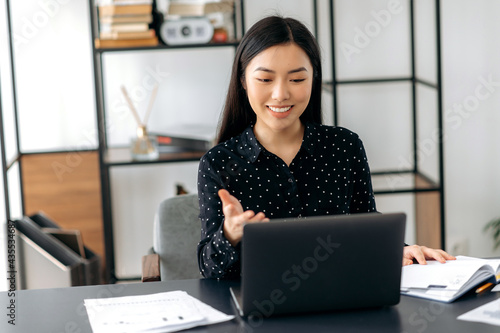Charismatic successful confident Asian business lady, freelancer, manager, using laptop, talking on video conference with client or colleague, discussing business project, gesturing with hand, smiling