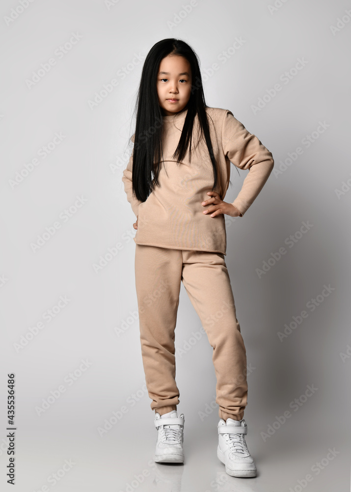 Cool Asian kid girl green pants, hoodie, sweater and sneakers stands holding hand at waist, posing in new sportswear, waiting and watching over gray background