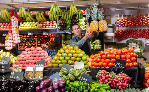 .Young male vendor in apron offering fresh fruits and vegetables on the supermarket  woman on background.