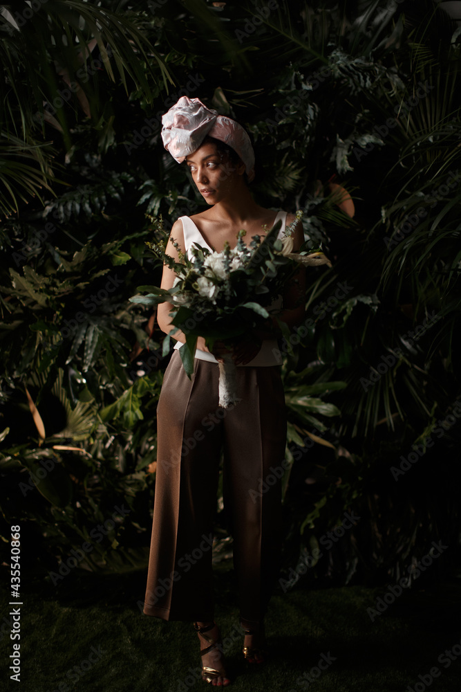 Charming calm curly hair, dark - skinned woman in white dress posing in a tropical decoration, holding a bouquet of colorful flowers in her hands Charming calm curly hair, dark - skinned woman in white dress posing in a tropical decoration, holding a bouquet of colorful flowers in her hands