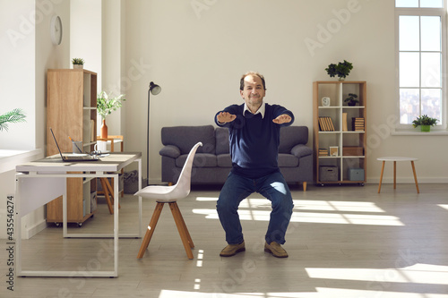 Senior man squatting in the living-room. Happy energetic mature businessman doing fitness warm up exercise during working day in home office. Concept of workplace workout for keeping fit and healthy