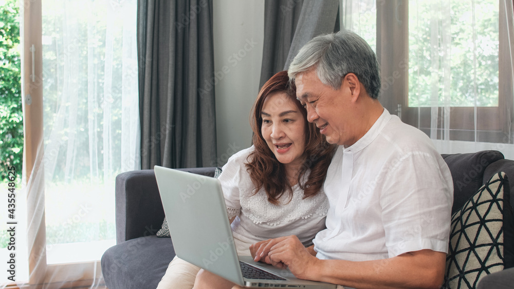 © tirachard - Asian senior couple using laptop at home. Asian Senior Chinese grandparents, surf the Internet to check social media while lying on sofa in living room at home concept.