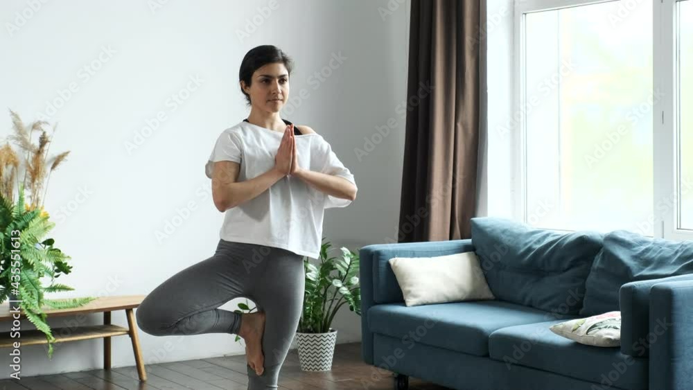 A Young Indian Woman Is Doing Morning Yoga, Doing The Exercise Standing on One Leg For Balance. Located at Home in a Bright Room A Young Indian Woman Is Doing Morning Yoga, Doing The Exercise Standing on One Leg For Balance. Located at Home in a Bright Room