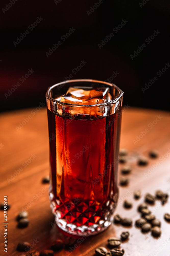 A cocktail in a collins highball glass with an ice spear, coffee beans around, on a wooden table, at the bar. A cocktail in a collins highball glass with an ice spear, coffee beans around, on a wooden table, at the bar.