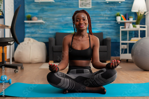 Portrait of black woman smiling practicing yoga at home sitting in lotus pose on mat mediting, practicing mindfulness meditation harmony, African american person doing relaxing workout. photo