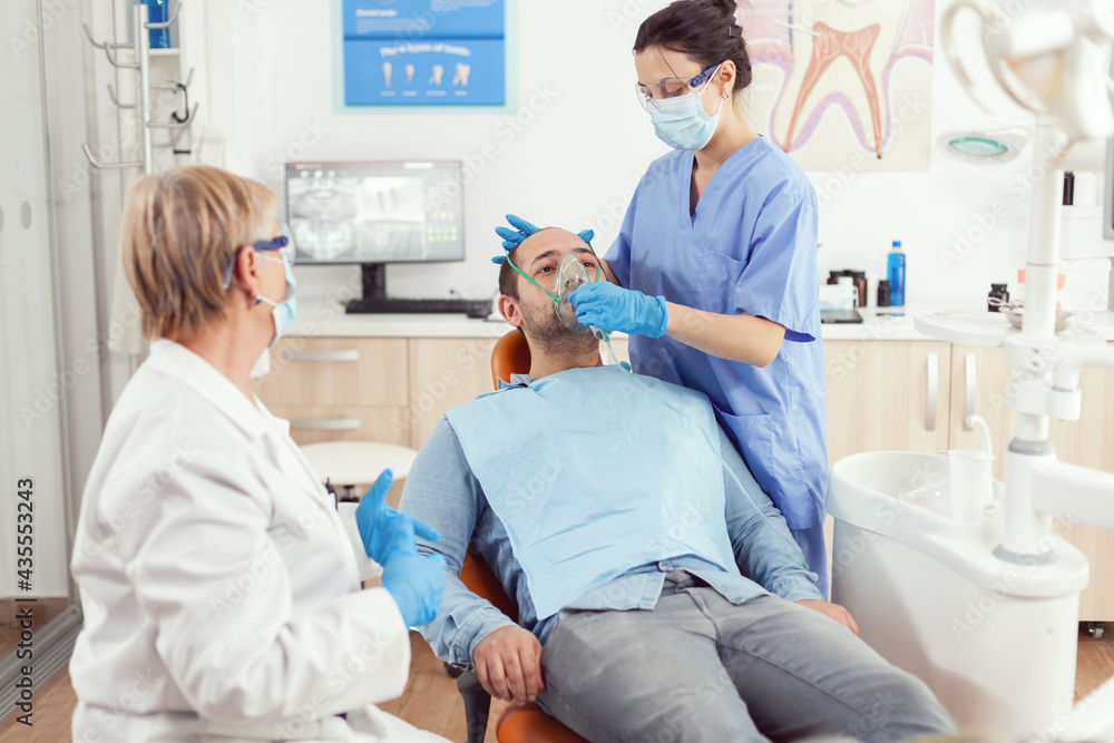 Stomatologist nurse putting oxigen mask before tooth surgery sitting on dental chair in stomatology cabinet. Senior doctor and nurse working in modern orthodontic office wearing mask and gloves Stomatologist nurse putting oxigen mask before tooth surgery sitting on dental chair in stomatology cabinet. Senior doctor and nurse working in modern orthodontic office wearing mask and gloves