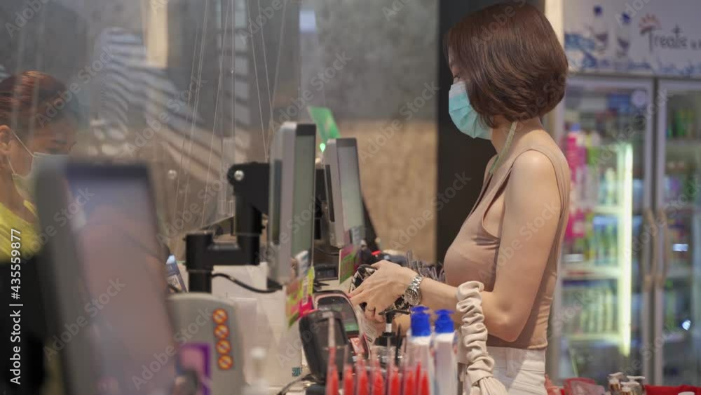 asian woman customer in medical face mask making payment at cashier with plexiglass at supermarket in department store. authentic shot during covid-19 and coronavirus pandemic outbreak. asian woman customer in medical face mask making payment at cashier with plexiglass at supermarket in department store. authentic shot during covid-19 and coronavirus pandemic outbreak.