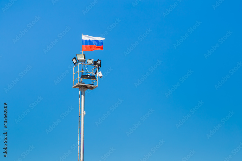 Lighting mast with Russian flag, CCTV camera and searchlights. Close-up against the clear blue sky. There is a copy space. The canvas of the flag develops smoothly in the wind. Lighting mast with Russian flag, CCTV camera and searchlights. Close-up against the clear blue sky. There is a copy space. The canvas of the flag develops smoothly in the wind.