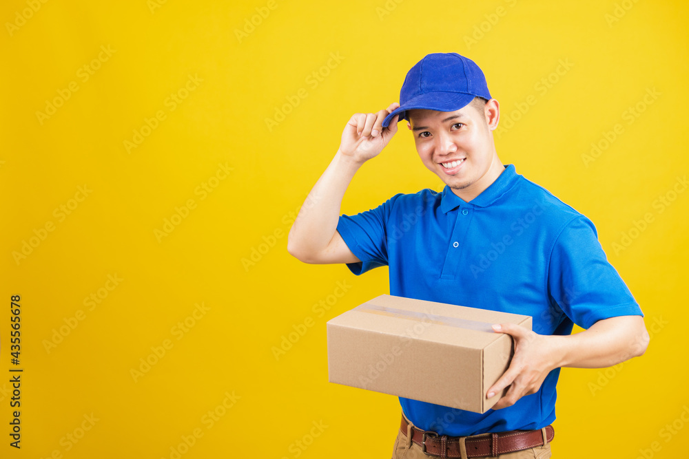 Portrait excited attractive delivery happy man logistic standing he smile wearing blue t-shirt and cap uniform holding parcel box looking to camera, studio shot isolated on yellow background Portrait excited attractive delivery happy man logistic standing he smile wearing blue t-shirt and cap uniform holding parcel box looking to camera, studio shot isolated on yellow background