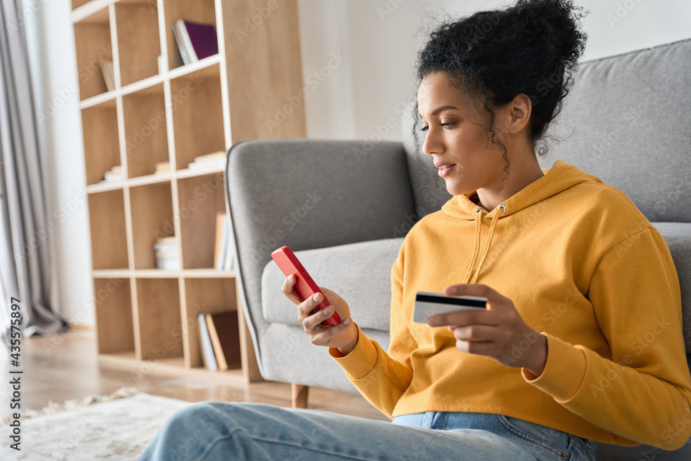 Young adult African American female consumer holding credit card and smartphone sitting on floor at home doing online banking transaction. E commerce virtual shopping, secure mobile banking concept. - obrazy, fototapety, plakaty 