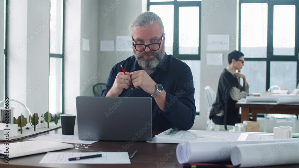 Portrait of confident senior male architect with grey hair and beard sitting at desk in office and looking at camera Portrait of confident senior male architect with grey hair and beard sitting at desk in office and looking at camera