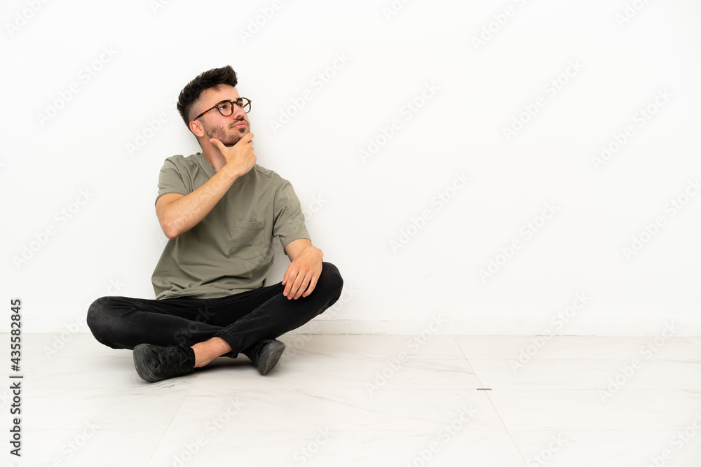 Young caucasian man sitting on the floor isolated on white background having doubts and with confuse face expression Young caucasian man sitting on the floor isolated on white background having doubts and with confuse face expression