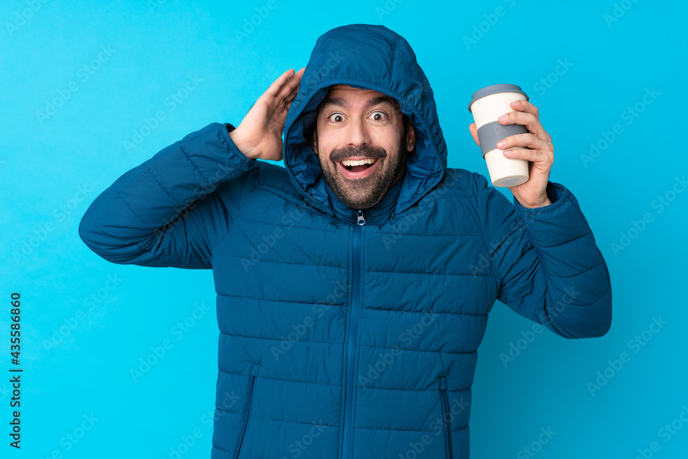 Naklejka premium Man wearing winter jacket and holding a takeaway coffee over isolated blue background with surprise expression