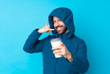 Man wearing winter jacket and holding a takeaway coffee over isolated blue background making phone gesture and pointing front