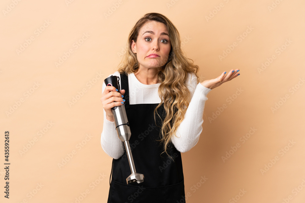 Young brazilian woman using hand blender isolated on beige background having doubts while raising hands Young brazilian woman using hand blender isolated on beige background having doubts while raising hands