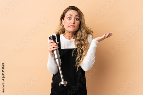 Young brazilian woman using hand blender isolated on beige background having doubts while raising hands