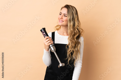 Young brazilian woman using hand blender isolated on beige background looking to the side and smiling