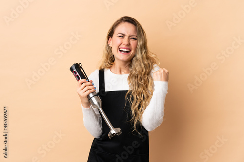 Young brazilian woman using hand blender isolated on beige background celebrating a victory in winner position