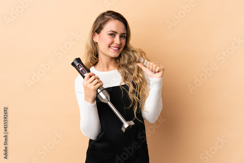 Young brazilian woman using hand blender isolated on beige background proud and self-satisfied