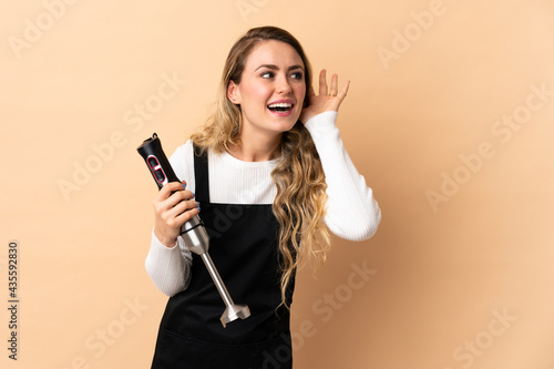 Young brazilian woman using hand blender isolated on beige background listening to something by putting hand on the ear