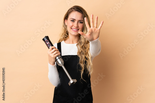 Young brazilian woman using hand blender isolated on beige background counting five with fingers