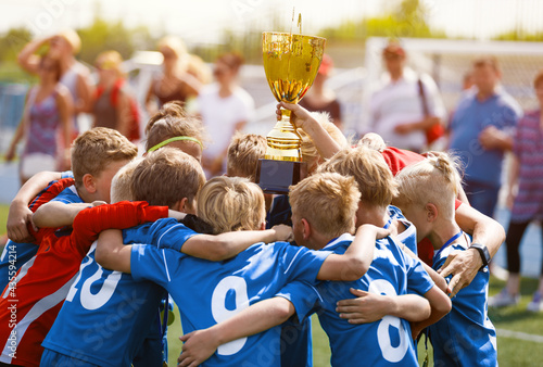 Fototapeta Naklejka Na Ścianę i Meble -  Happy Children in Football Team Winning Golden Cup in School Tournament. Young Boys in Blue Soccer Uniforms Rising Up Golden Cup. Young Coach Celebrating Success With Kids Football Players