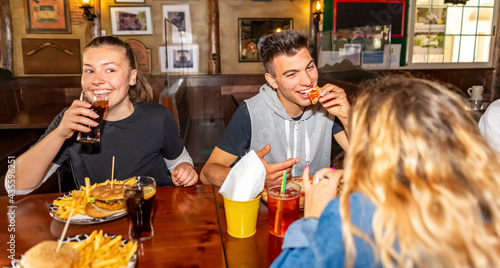 Group of millennials friends eating at fast food indoors in irish pub restaurant. Happy people partying and eating together. Young active teenagers with burgers, pizza and drinks having fun dining.