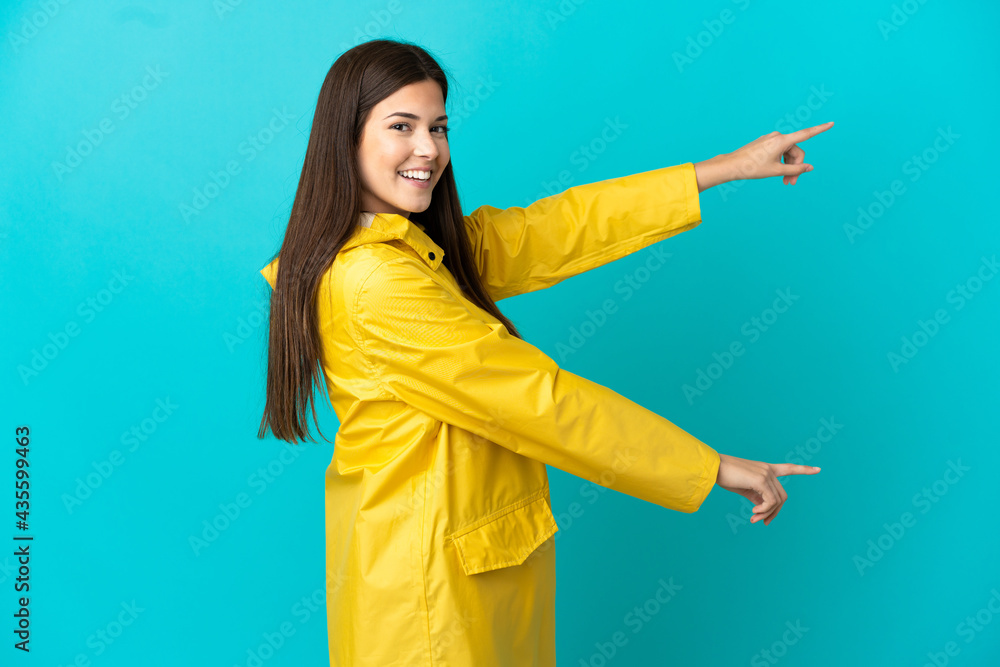 Teenager Brazilian girl wearing a rainproof coat over isolated blue background pointing finger to the side and presenting a product Teenager Brazilian girl wearing a rainproof coat over isolated blue background pointing finger to the side and presenting a product
