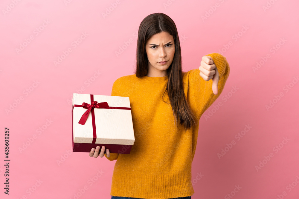 Teenager Brazilian girl holding a gift over isolated pink background showing thumb down with negative expression Teenager Brazilian girl holding a gift over isolated pink background showing thumb down with negative expression