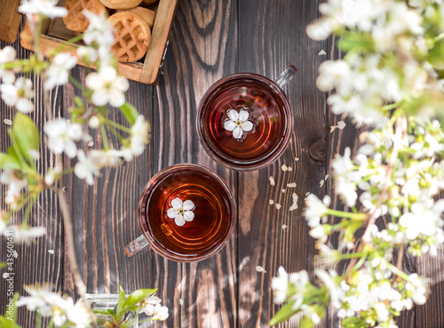 Tea party on a wooden table with branches of blossoming cherry. Black tea in glass mugs. White cherry flowers in a mug. Wooden chest with cookies on the table. View from above. Flatlay