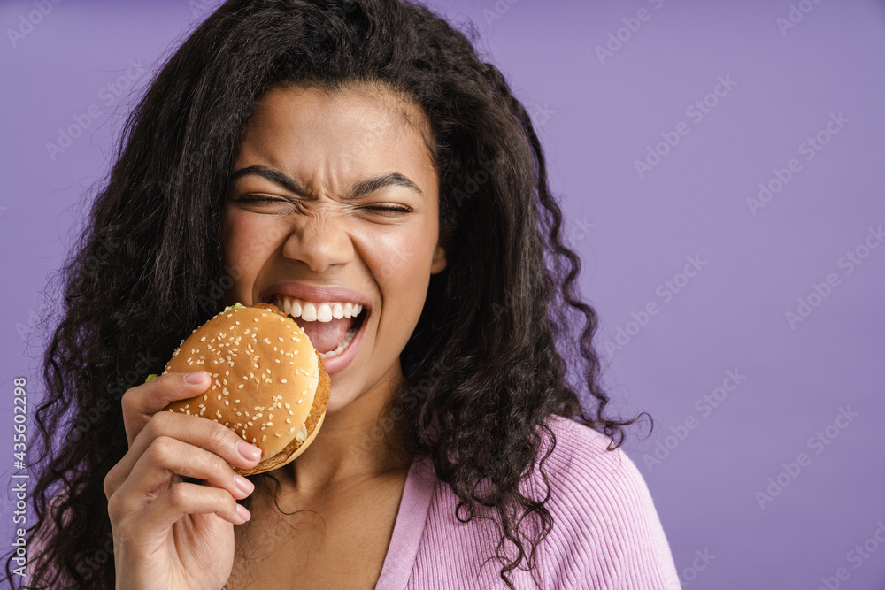 Young black woman with curly hair grimacing while eating hamburger