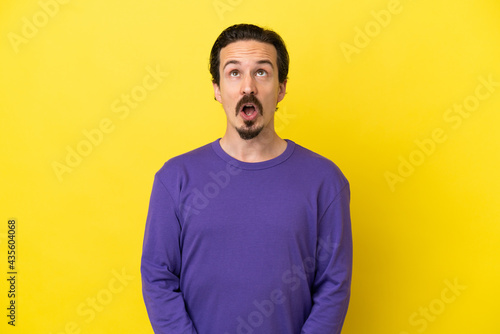 Young caucasian man isolated on yellow background looking up and with surprised expression