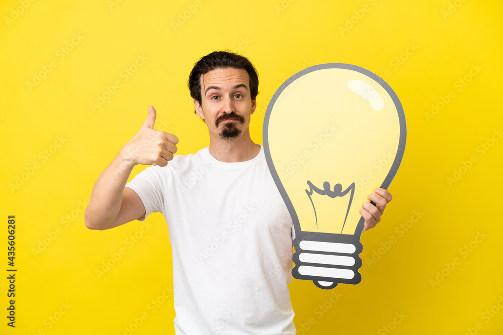 Young caucasian man isolated on yellow background holding a bulb icon with thumb up Young caucasian man isolated on yellow background holding a bulb icon with thumb up