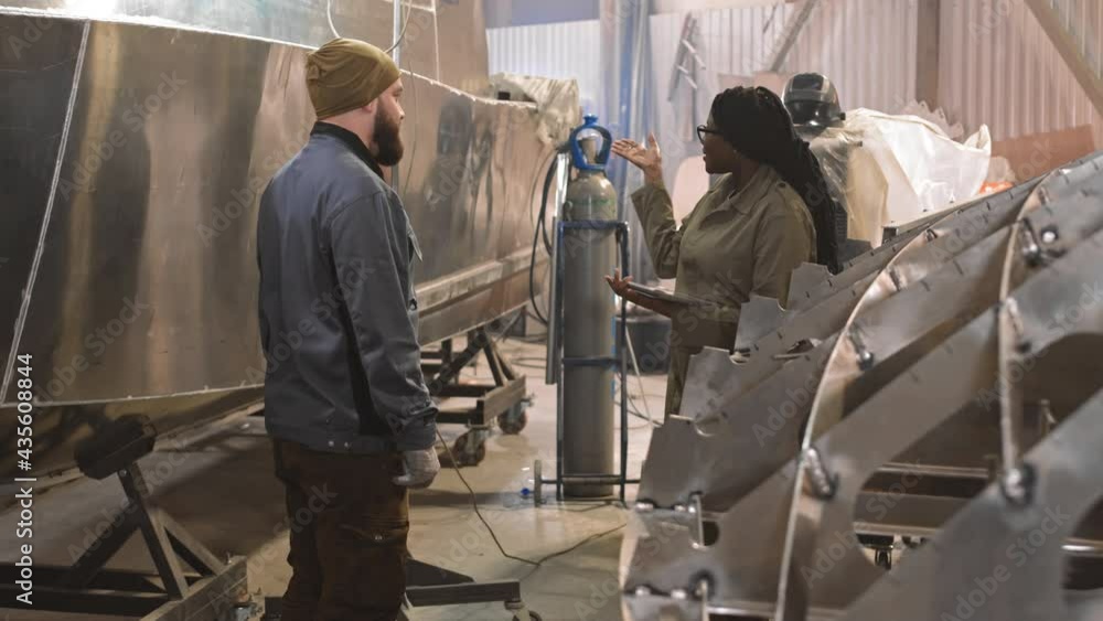 Medium long of bearded man standing in indoor dockyard, nearby metallic boats in progress, listening to young African female engineer wearing worker clothing, holding tablet computer and talking Medium long of bearded man standing in indoor dockyard, nearby metallic boats in progress, listening to young African female engineer wearing worker clothing, holding tablet computer and talking