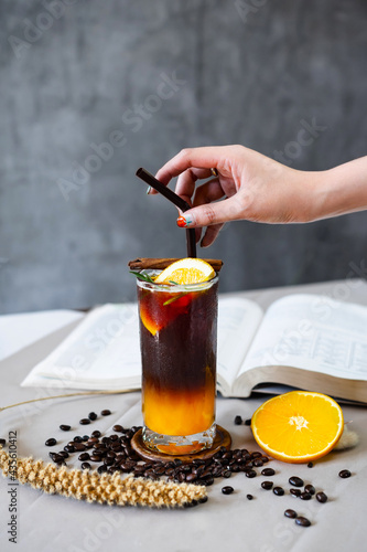 Orange black coffee and open book on the coffee table surrounded with coffee bean, hand touching a straw.