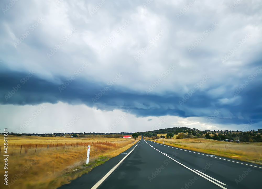 View of straight country road in Australia with flat surrounding farmland and threatening storm clouds and rain in the background.
