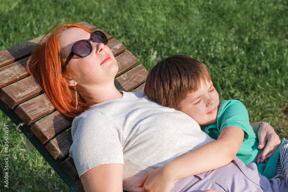 Young woman with red hair and boy are lie in park on wooden deck chair. Son hugs his mother gently. Happy parenting concept, relationship with children. Attachment of young children to their mother. Young woman with red hair and boy are lie in park on wooden deck chair. Son hugs his mother gently. Happy parenting concept, relationship with children. Attachment of young children to their mother.