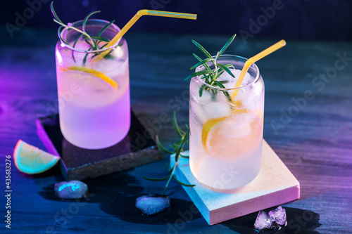 Two glasses of alcoholic cocktails with gin and lemonade , lemon and rosemary. Black wooden table, stone stands. Neon stylish back light