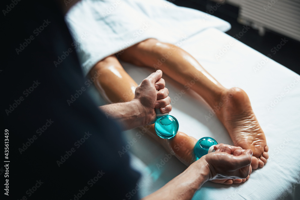 Charming young woman lying on massage table while having manual massage with cold glass in wellness center