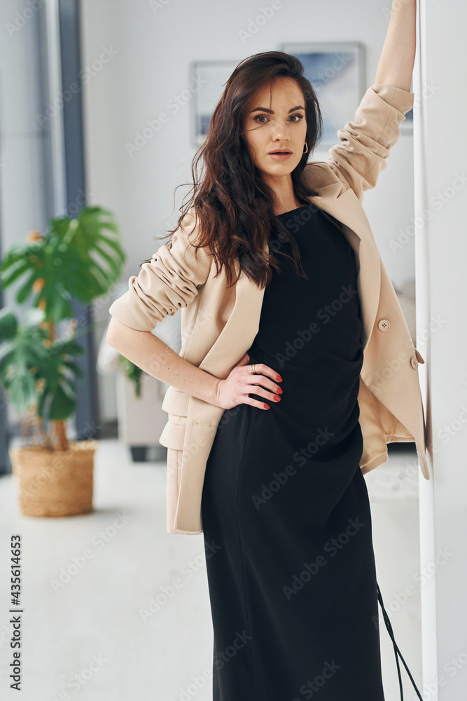 In black dress and jacket. European woman in fashionable stylish clothes is posing indoors