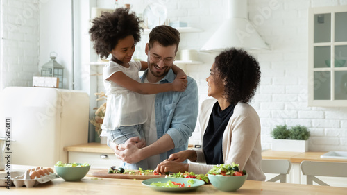 Smiling young multiracial mom and dad cook healthy delicious vegetarian salad with ethnic small daughter. Happy multiethnic family with girl child have fun prepare food in kitchen. Adoption concept.