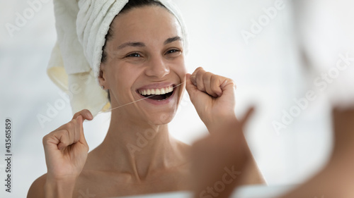 Happy young woman wrapped in bath towel flossing white healthy teeth with thread after shower  looking at reflection in mirror  smiling with open mouth. Dental care  oral hygiene  routine concept