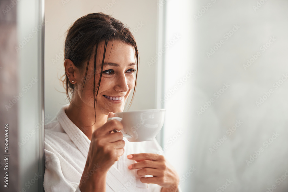 Laughing beautiful elegant lady in white bathrobe looking away while holding cup in hand in room indoors