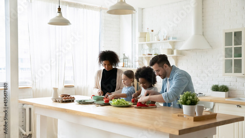 Young multiethnic family with small diverse teen daughters prepare healthy tasty food in kitchen together. Multiracial mom and dad cook at home with little girls children. Adoption, custody concept.