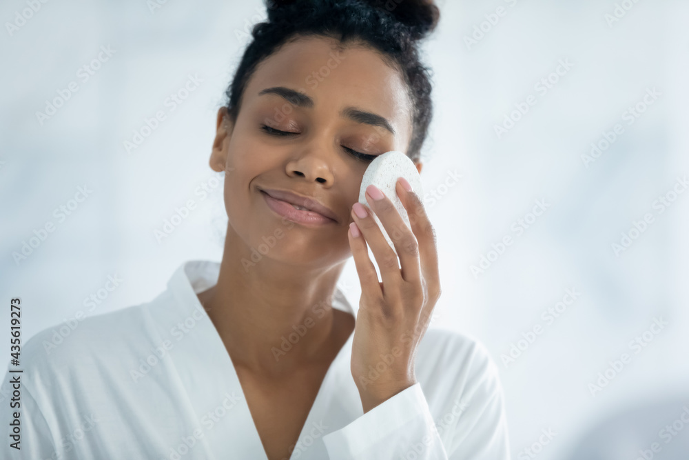 Happy relaxed Black girl cleaning facial skin from makeup, cosmetics with sponge at mirror, applying cleansing lotion with closed eyes, enjoying beauty care routine. Skincare concept. Head shot