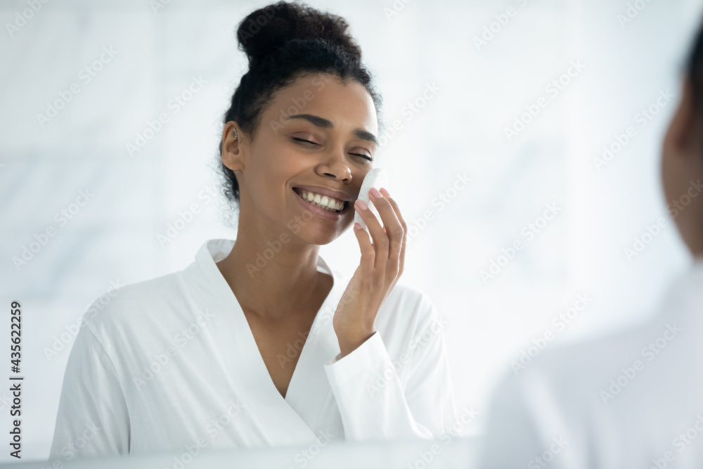 Happy young African American woman enjoying home spa skincare detox procedures, cleaning face with lotion and exfoliating sponge for fresh clear smooth skin, removing cosmetics and makeup Happy young African American woman enjoying home spa skincare detox procedures, cleaning face with lotion and exfoliating sponge for fresh clear smooth skin, removing cosmetics and makeup