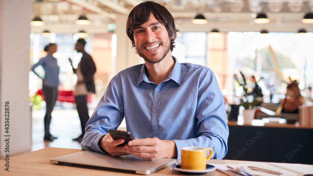 Portrait Of Smiling Businessman Sitting At Desk With Laptop In Modern Open Plan Office Portrait Of Smiling Businessman Sitting At Desk With Laptop In Modern Open Plan Office