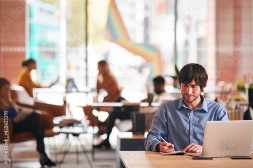 Businessman Sitting At Desk Writing In Notebook In Modern Open Plan Office